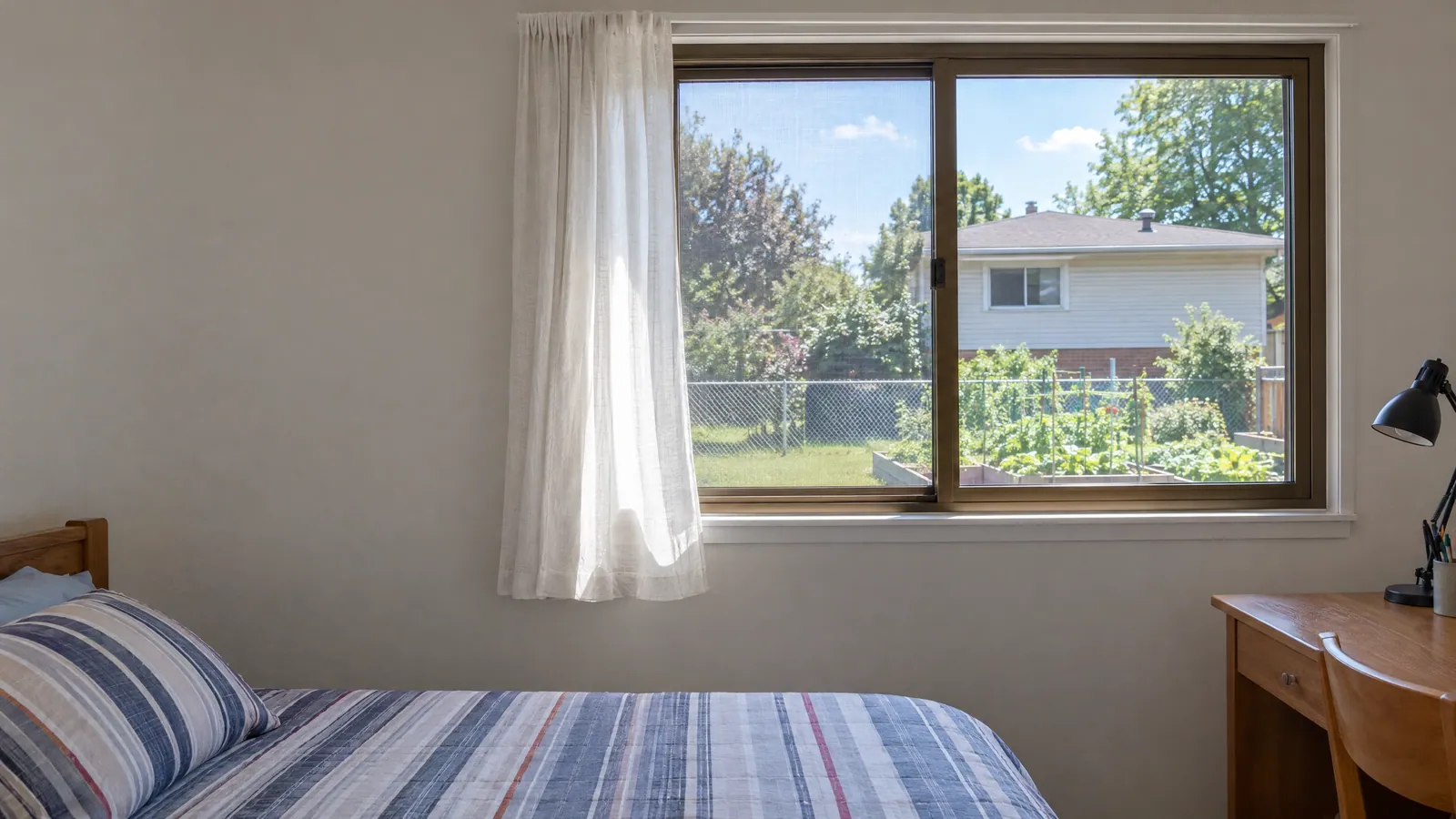 Bronze-coloured aluminum horizontal slider window partly open in a 1970s bedroom, looking out on a sunny backyard with a chain-link fence