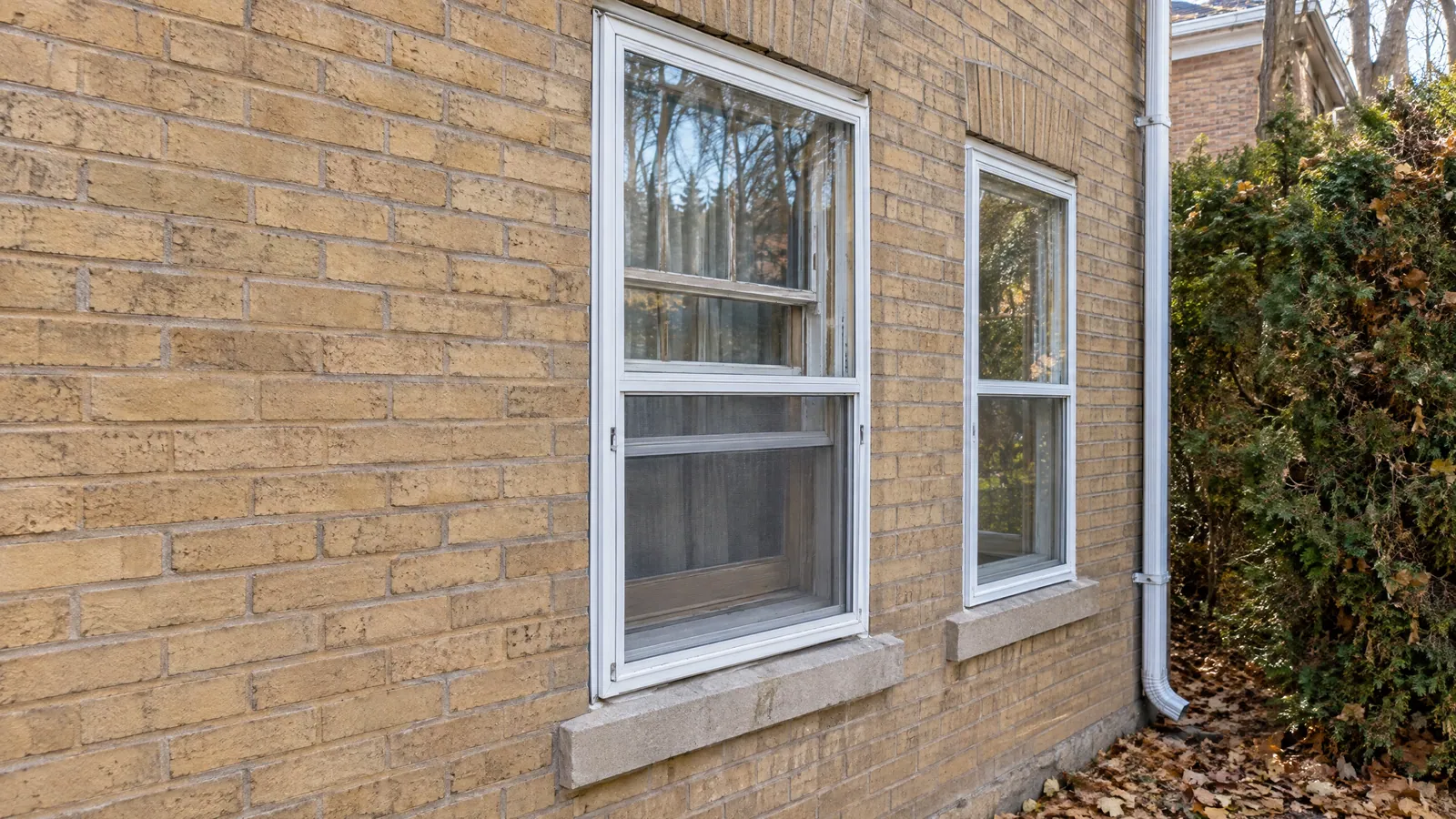 White aluminum triple-track storm window fitted over an original wood double-hung sash on a buff-brick house, with fallen leaves on the ground below