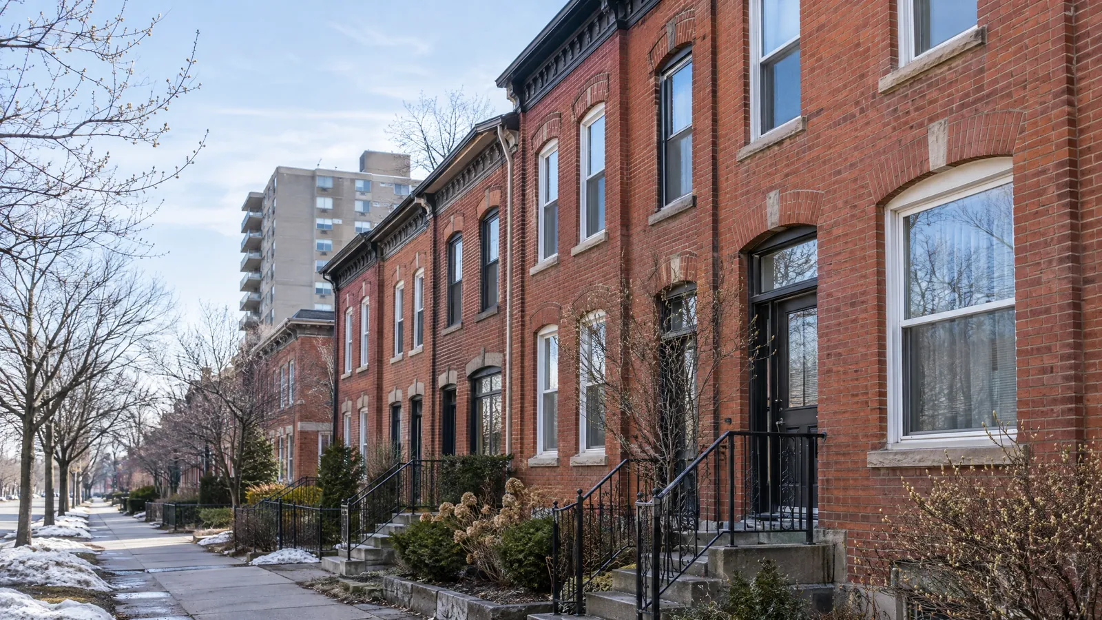Row of 19th-century red-brick rowhouses with tall arched windows and iron stair railings, an apartment tower behind, in early spring