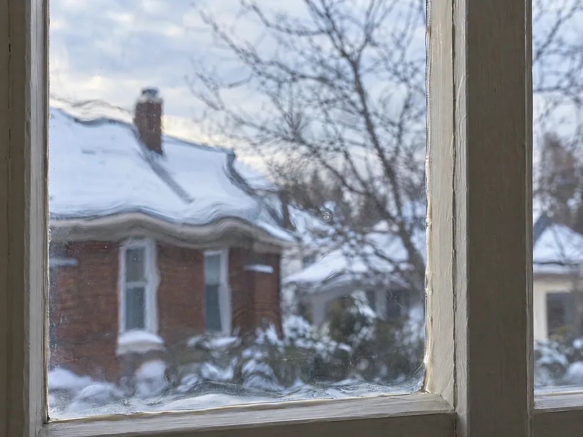 Close-up of old window glass with a slight wave and a small air bubble, bending the view of a snowy roofline beyond a painted wood muntin