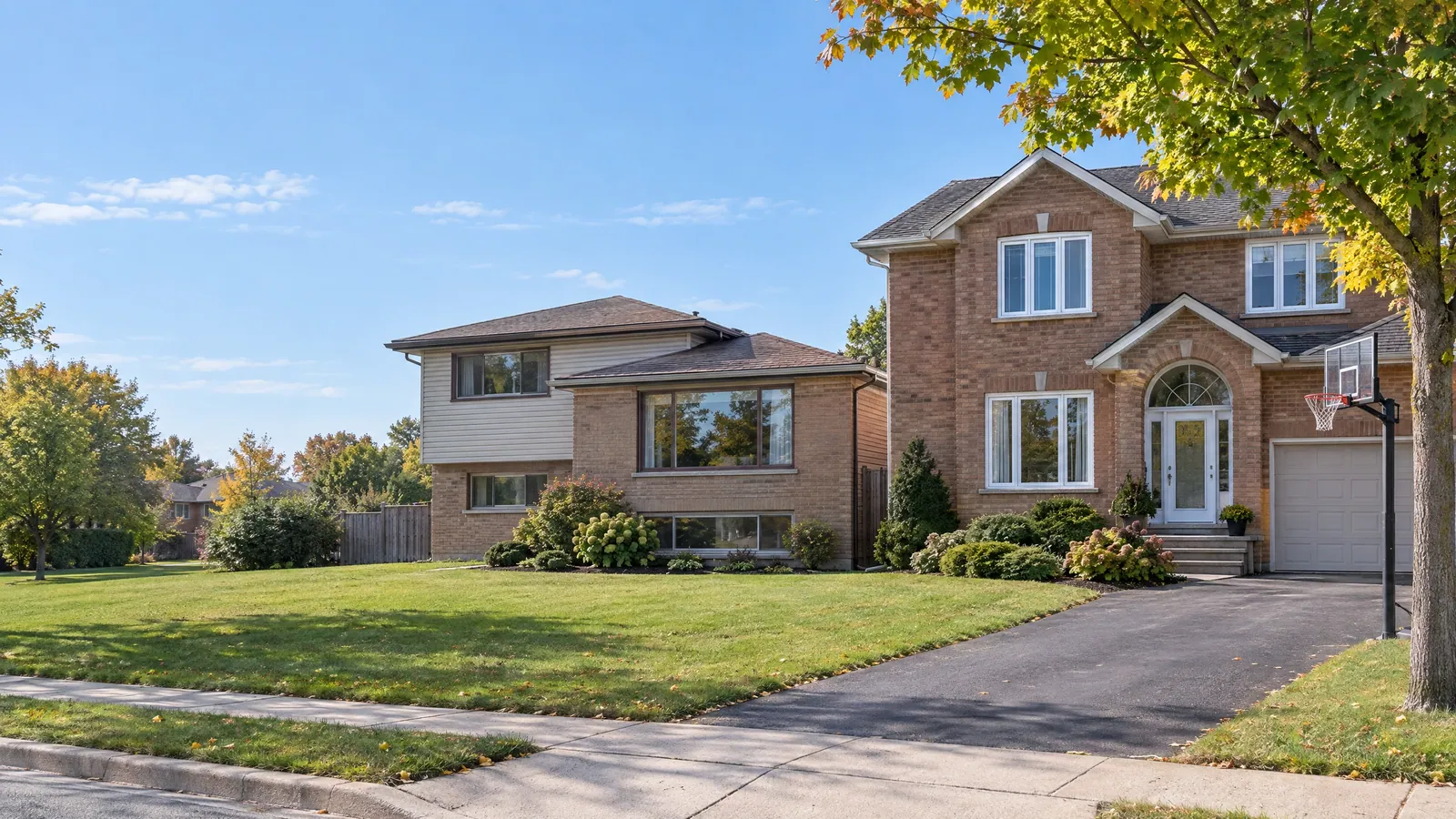 A 1970s brick back-split with aluminum sliding windows next to a 1990s two-storey house with vinyl casement windows, in early autumn